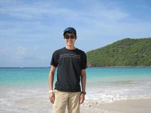 Man in black t-shirt with blue hat and sunglassess on beach with blue water and green hill behind