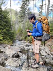 Asian man in blue looking at phone with orange backpack in woods