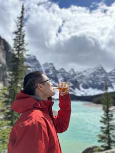 Asian man in red jacket drinking bottle of maple syrup in front of lake and mountains