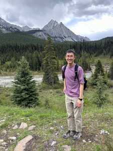 Smiling Asian man in purple shirt in front of stream and mountains on a cloudy day