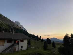 Concrete hut at dusk in front of mountain with moon above and grassy field with trees to the right